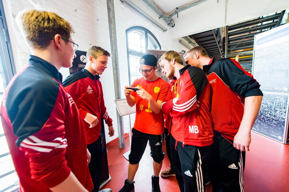 Sechs Personen in Sportbekleidung stehen zusammen und schauen auf ein Smartphone in einer hellen Umgebung.