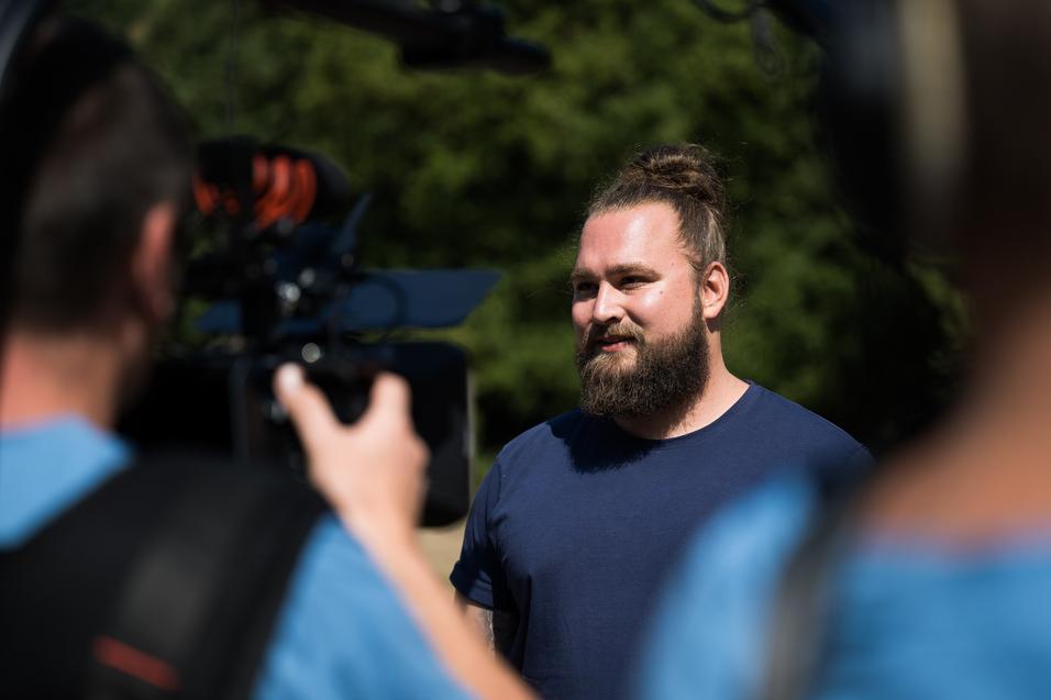 Mann mit Bart und Dutt steht vor einer Kamera und spricht, während ein weiteres Kamerateam im Hintergrund sichtbar ist.