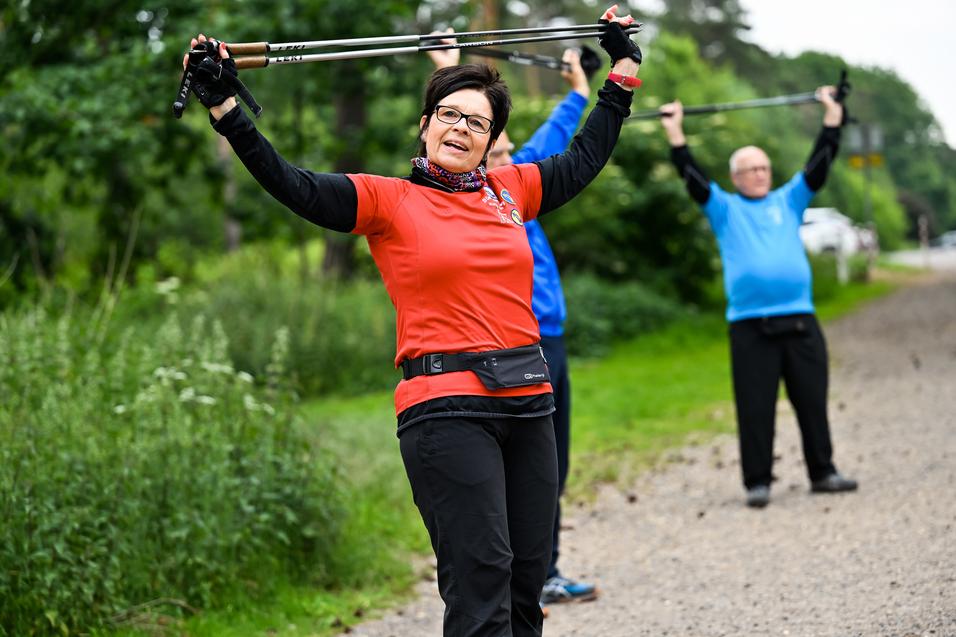 Frau in rotem Shirt hebt Wanderstöcke über den Kopf, während im Hintergrund zwei Personen in blauer und schwarzer Kleidung trainieren.